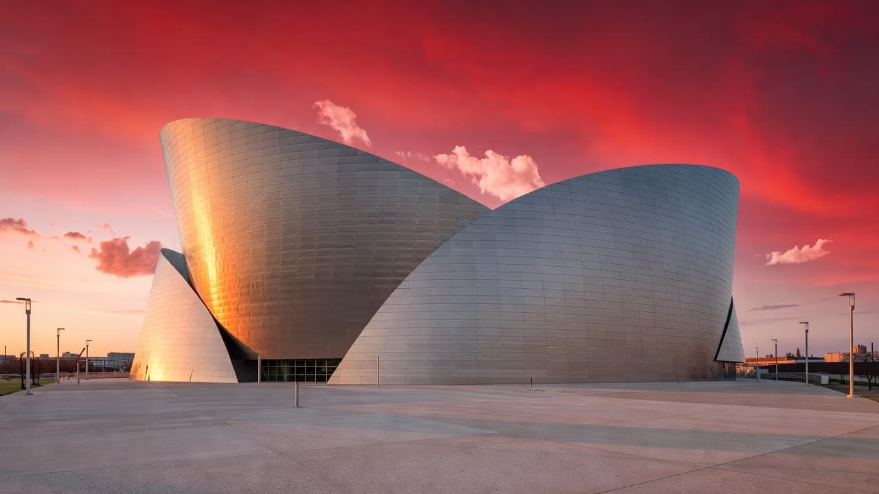 The white glass shells of the Kauffman Center rise against a vivid sunset sky saturated with deep crimson and orange hues. The building exterior glows softly while reflecting the dramatic evening light.