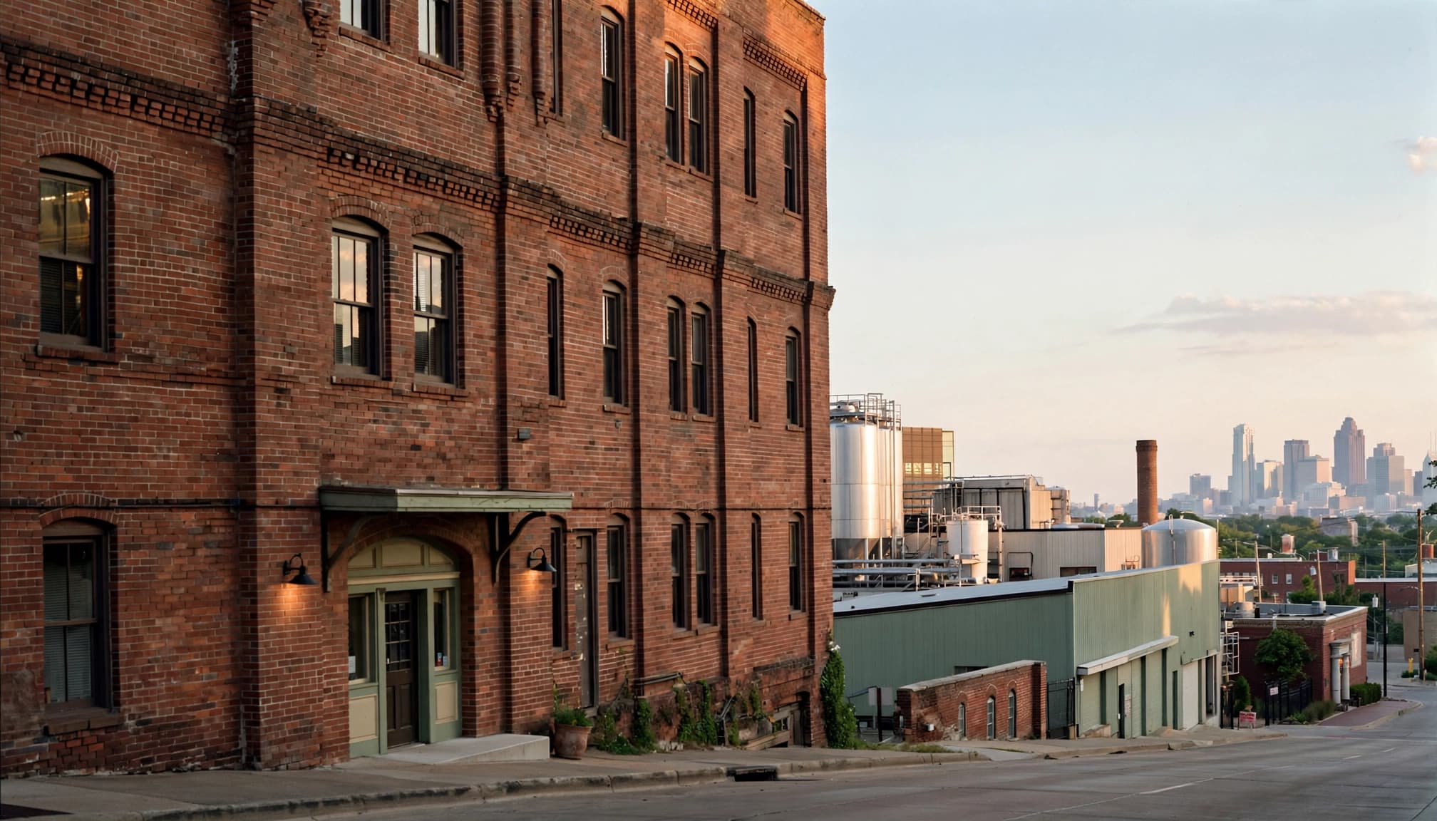 Brick brewery buildings beside a sidewalk, with the downtown skyline in the distance.